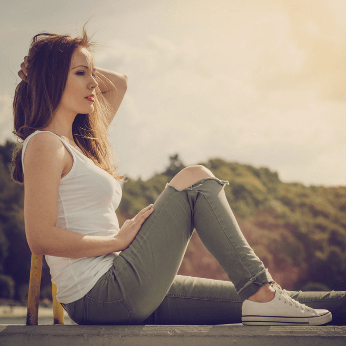Young woman sitting outdoors in casual outfit, enjoying a sunny day with nature in the background.