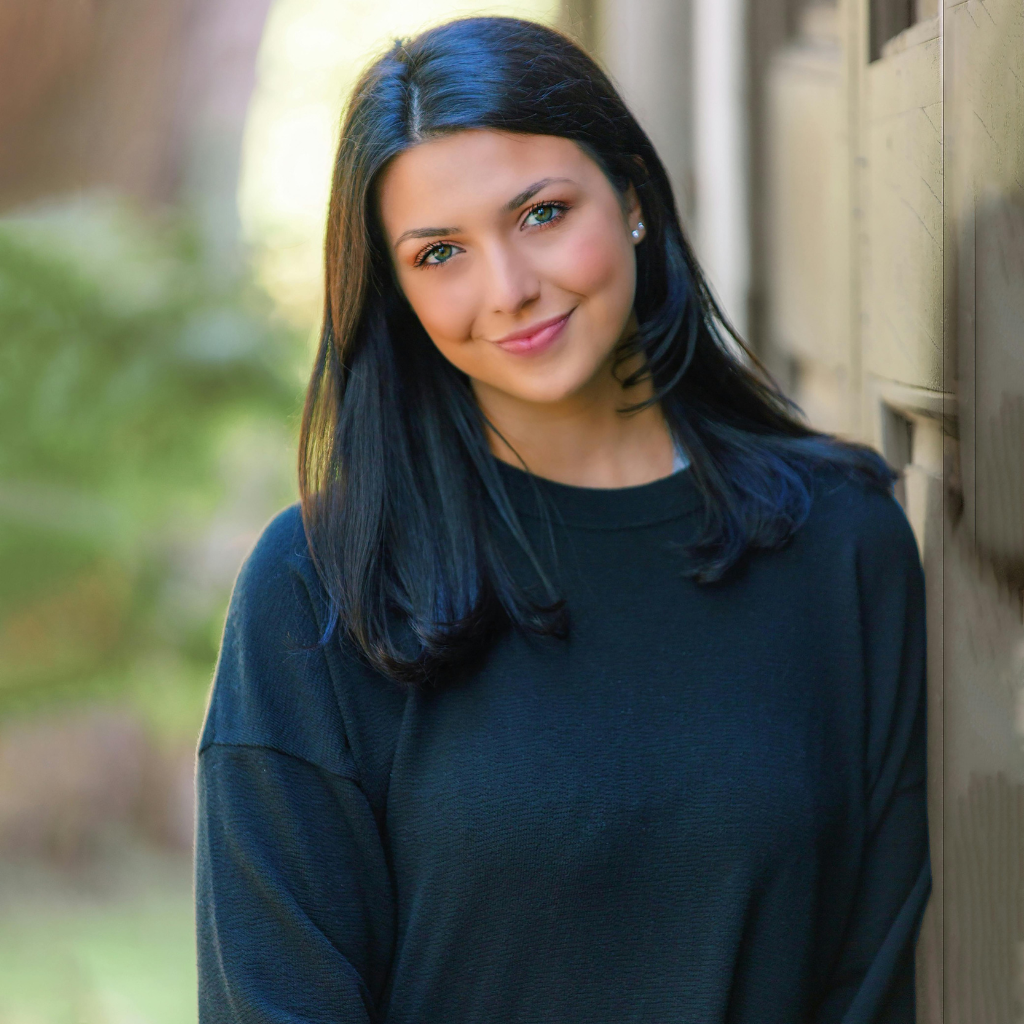 A young woman with long dark hair smiling while wearing a black sweater with a natural background.