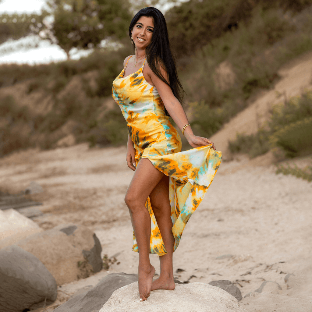 Woman in a vibrant yellow and blue dress twirling on a rocky beach, enjoying a sunny day outdoors.