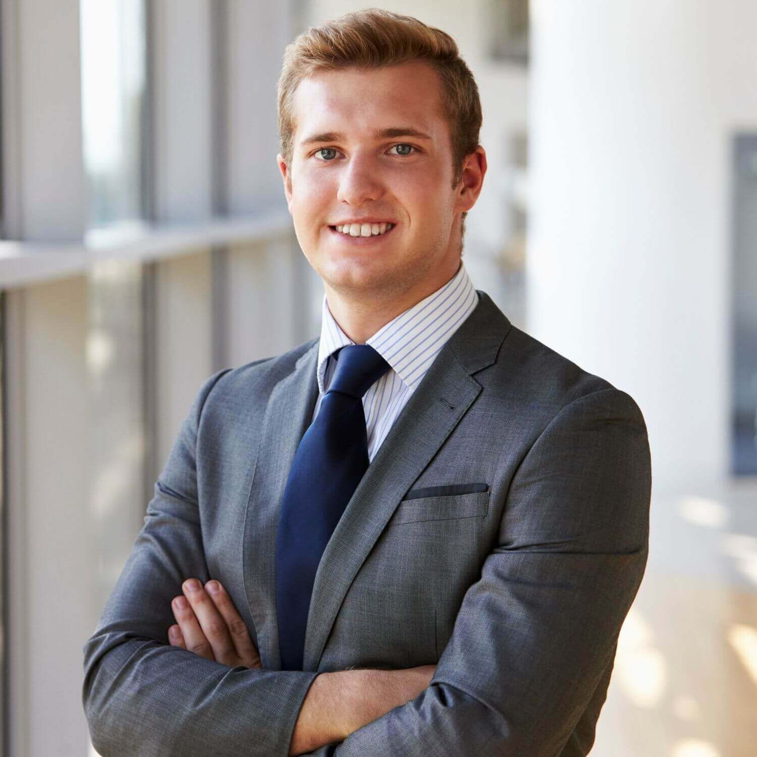 Smiling professional man in a suit, standing confidently with arms crossed, against a modern office background.